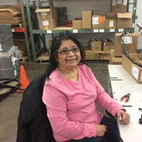 Sophia sits at a work desk with some tools and shelves with boxes behind her. She is wearing a pinks shirt and dark glasses with a friendly smile.
