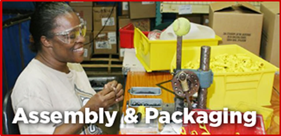 A smiling woman sits at a workbench that has several bins of parts. She is wearing safety glasses and putting some components together. 