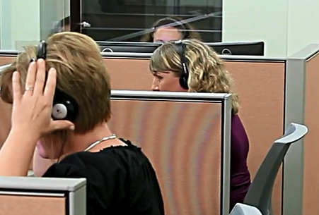A row of cubicles with people seated and wearing telephone headphones. 