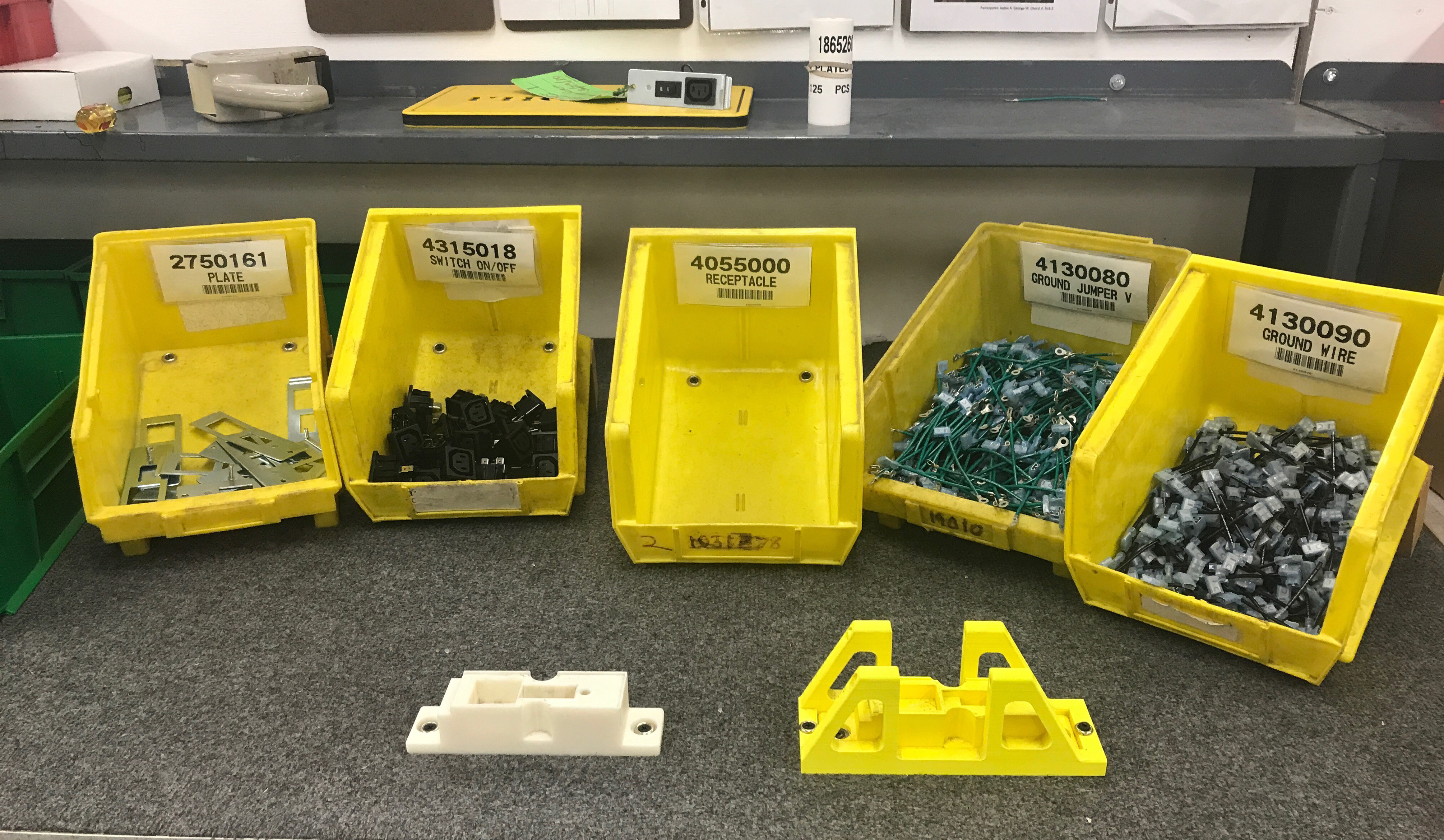 A work station has a series of five yellow bins, each containing different parts. In front of them are two small plastic jigs. 